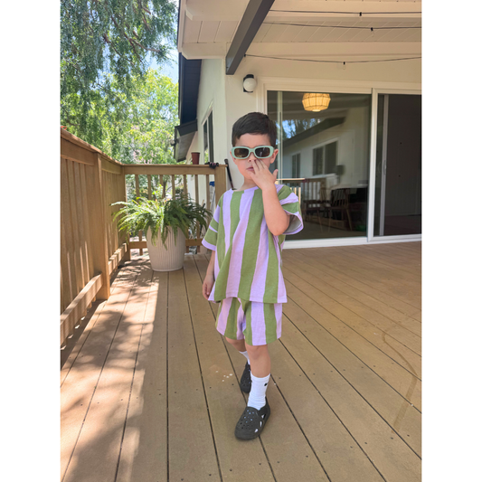 Green/Purple | A young child in sunglasses makes a peace sign while wearing the UMBRELLA STRIPE SHORT SET, a green-and-lavender striped outfit made from organic cotton, standing on a wooden deck.