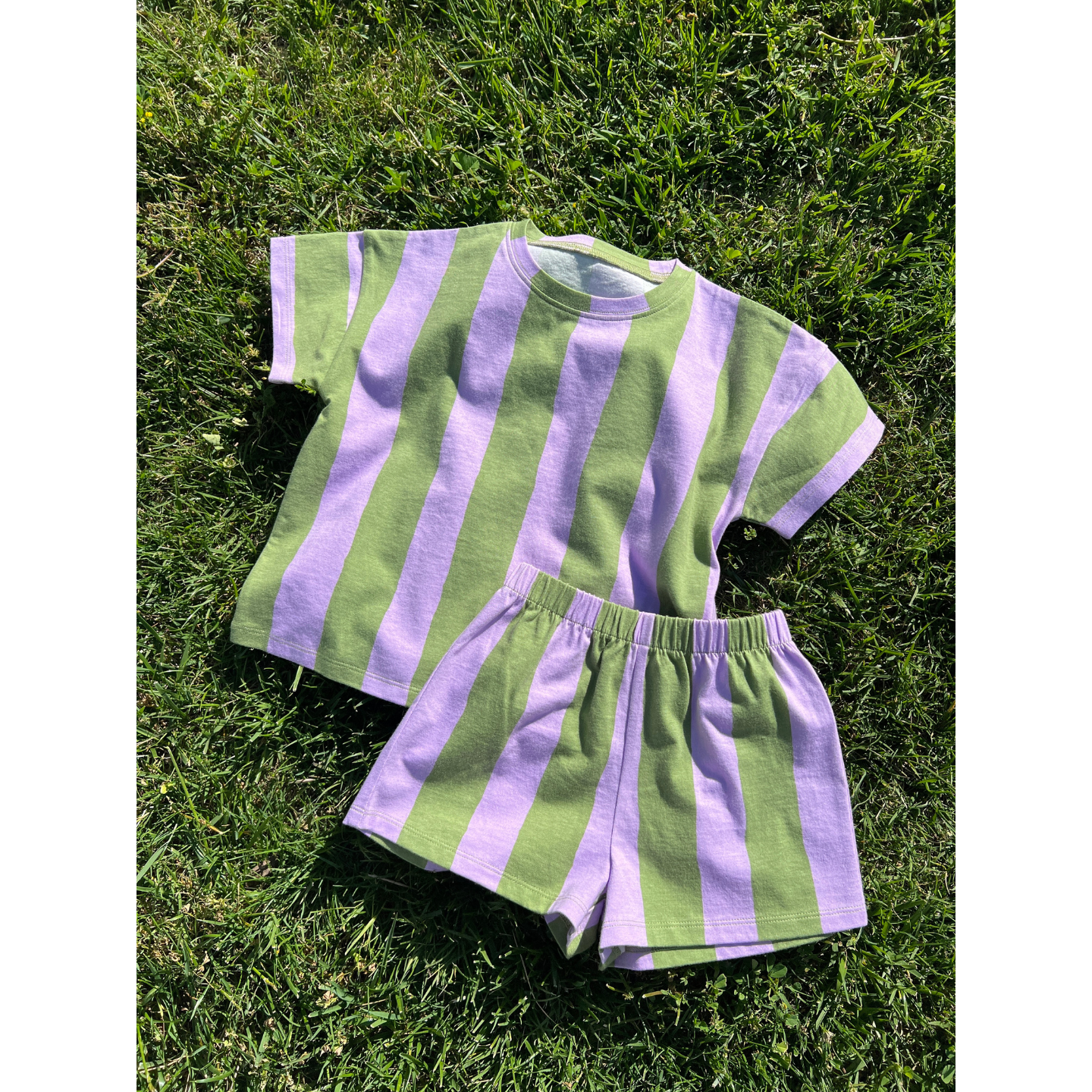Green/Purple | The UMBRELLA STRIPE SHORT SET, a unisex short-sleeve shirt and shorts with bold green and lavender stripes, is displayed on grass—ideal for playful days in stylish striped sets.