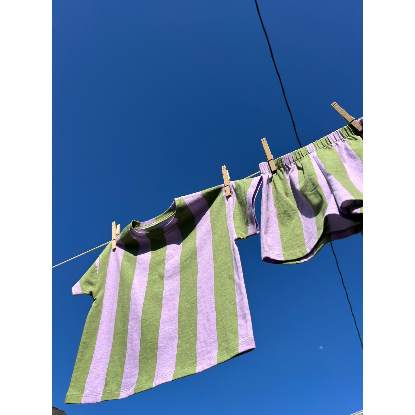 Green/Purple | The UMBRELLA STRIPE SHORT SET—a green and lavender striped organic cotton t-shirt and matching shorts—hangs on a clothesline with clothespins under a clear blue sky.