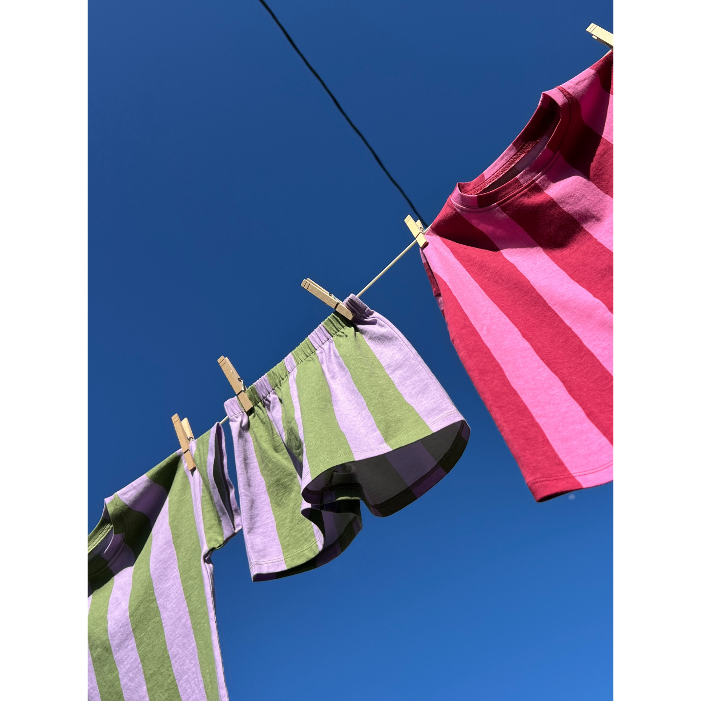 Green/Purple | The UMBRELLA STRIPE SHORT SET, made from organic cotton in purple, green, and red stripes, hangs with wooden clothespins on a clothesline beneath a clear blue sky.