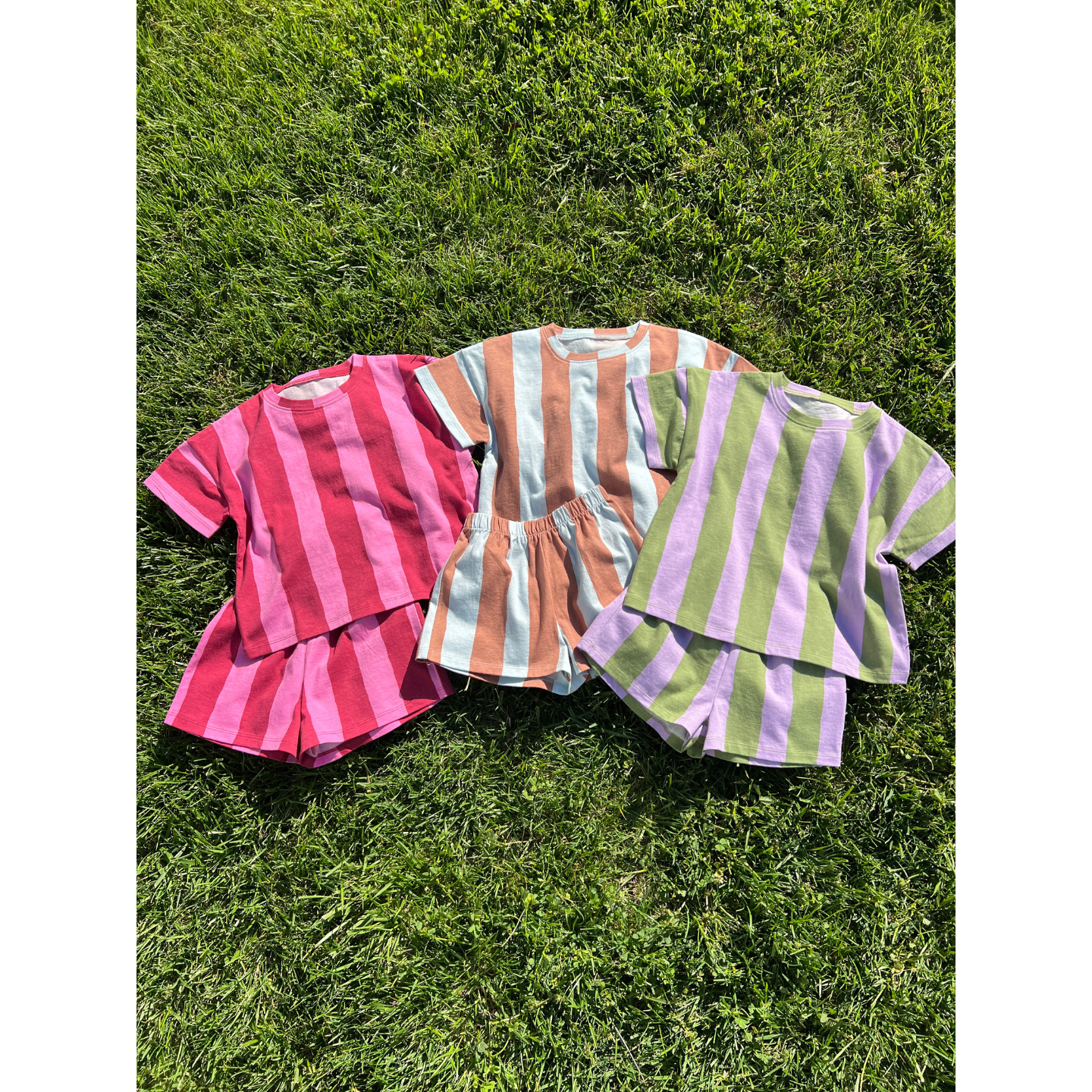 Blue/Brown | Three unisex UMBRELLA STRIPE SHORT SETS in red-pink, orange-blue, and green-purple striped colorways are displayed on green grass. Each set is made from soft, breathable organic cotton for all-day comfort.