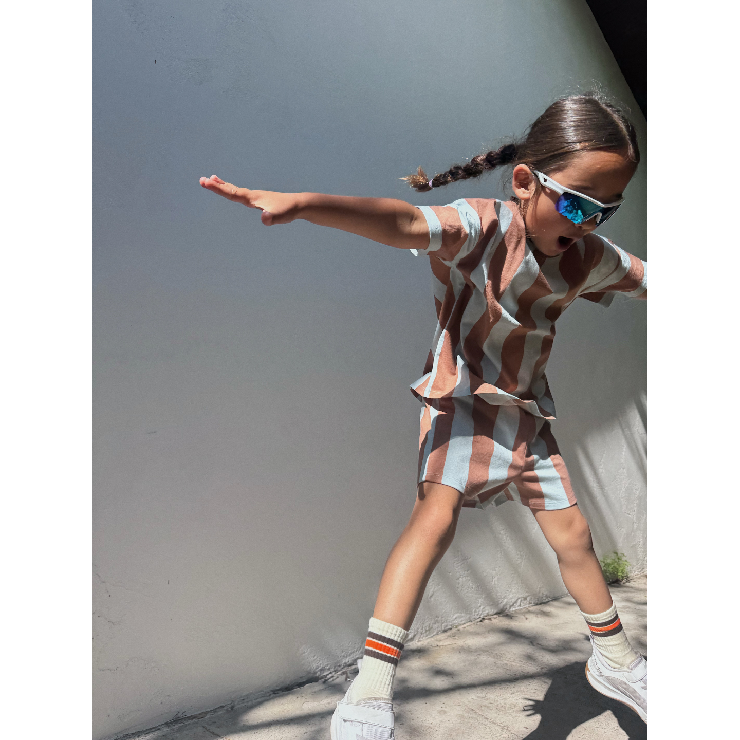 Blue/Brown | A child in the UMBRELLA STRIPE SHORT SET, sunglasses, and sneakers jumps with arms outstretched against a plain wall in sunlight.