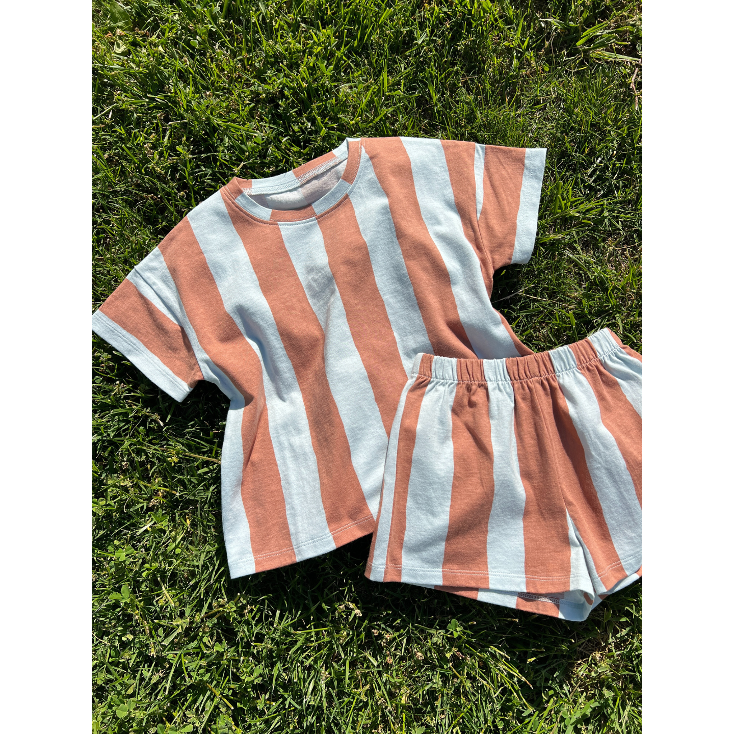 Blue/Brown | The UMBRELLA STRIPE SHORT SET, made from organic cotton, is a unisex set with a short-sleeve shirt and shorts in bold white and rust-orange stripes, pictured on green grass.