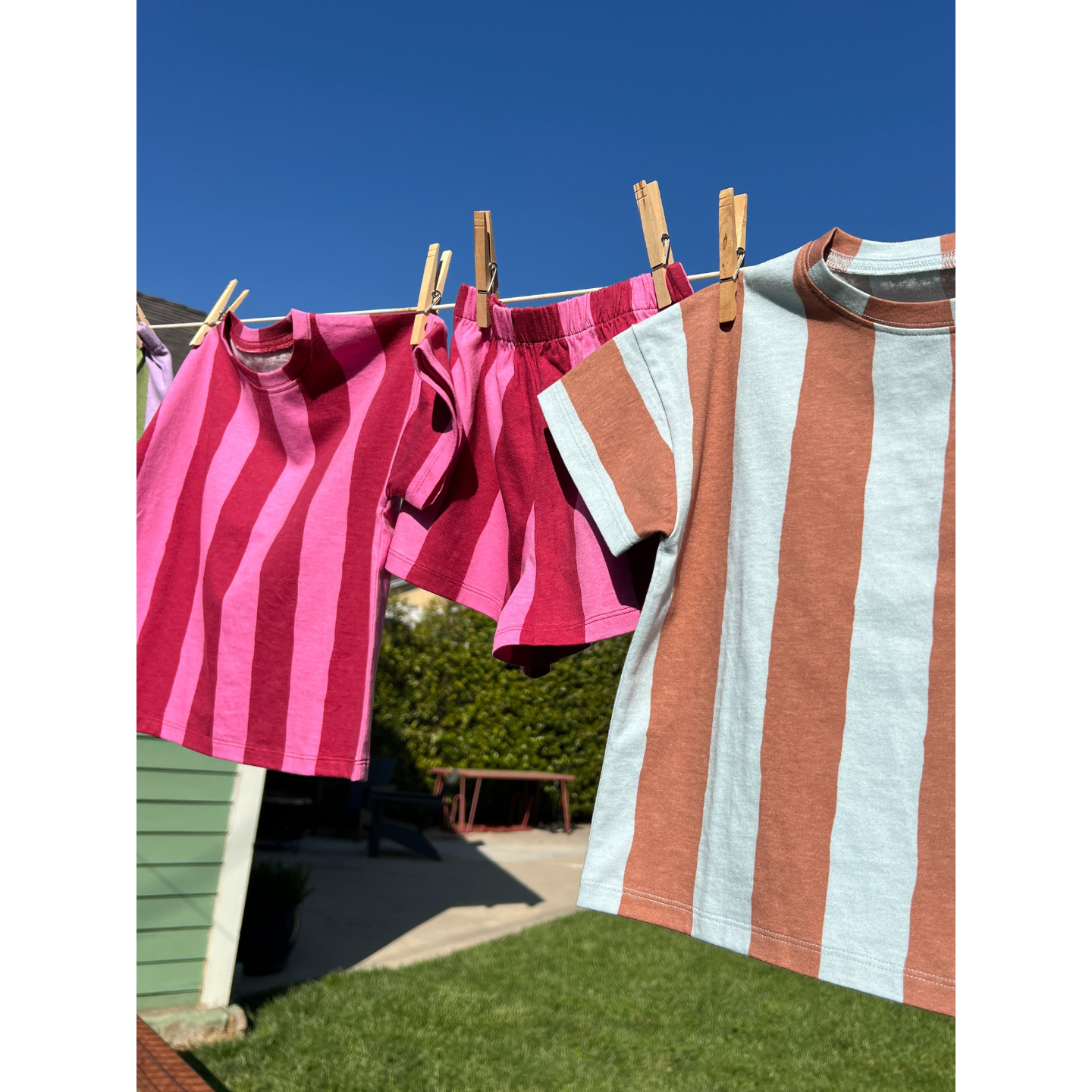 Blue/Brown | Three UMBRELLA STRIPE SHORT SETS—made from organic cotton, in two pink and one brown and blue—hang on a clothesline outside under a clear blue sky.