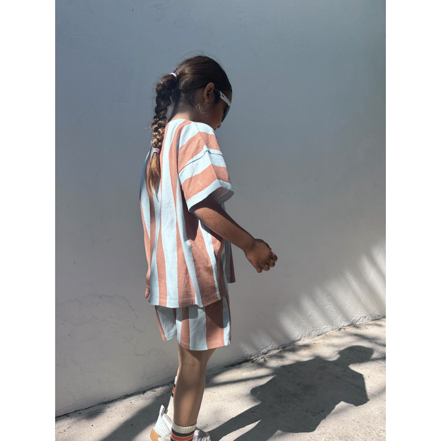 Blue/Brown | A young girl with braided hair and sunglasses wears the UMBRELLA STRIPE SHORT SET, made from organic cotton, as she stands in sunlight by a white wall.