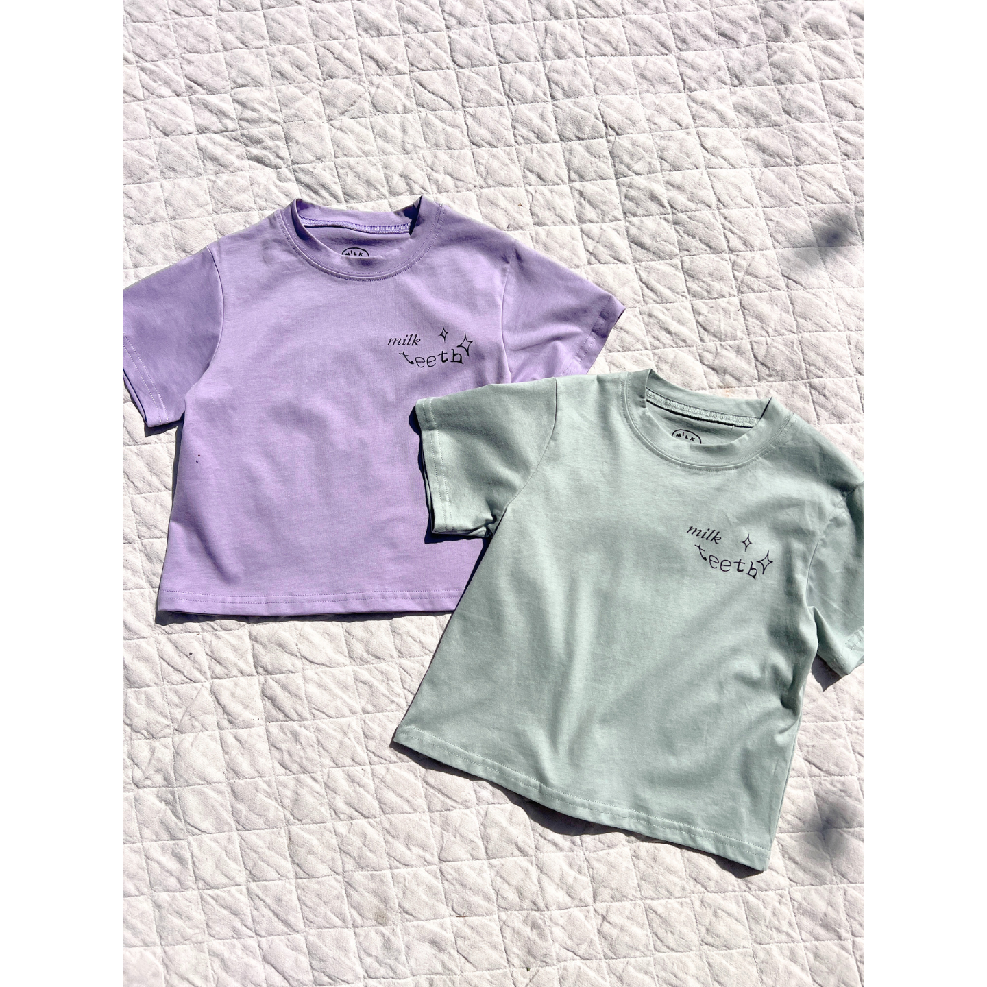 Mint | Two MILK TEETH TEE shirts in lavender and mint green, featuring "milk teeth" and star designs by artists, are laid flat on a quilted white surface.