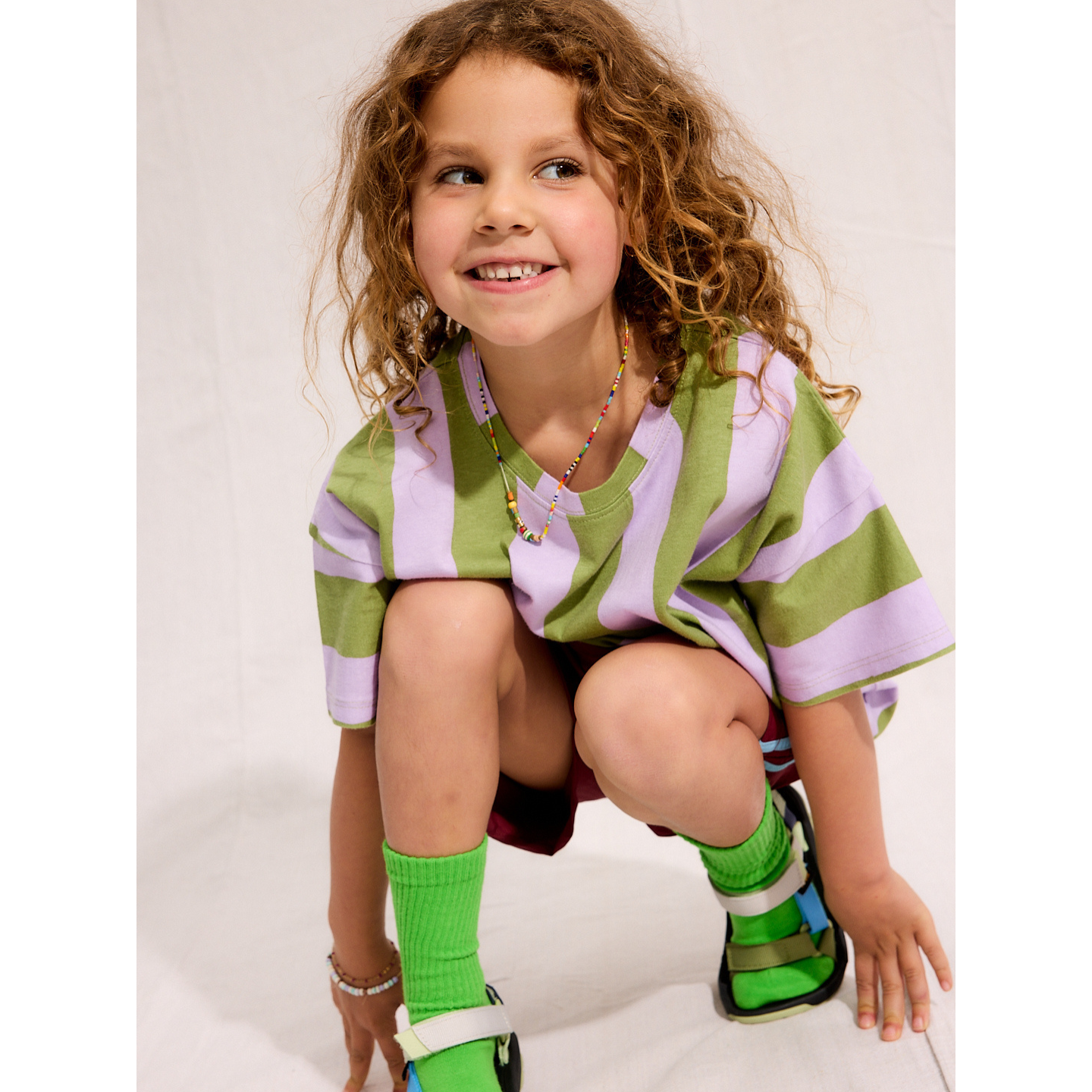 Green/Purple | A child is wearing the Umbrella Stripe Set with green socks and sandals while squatting and smiling