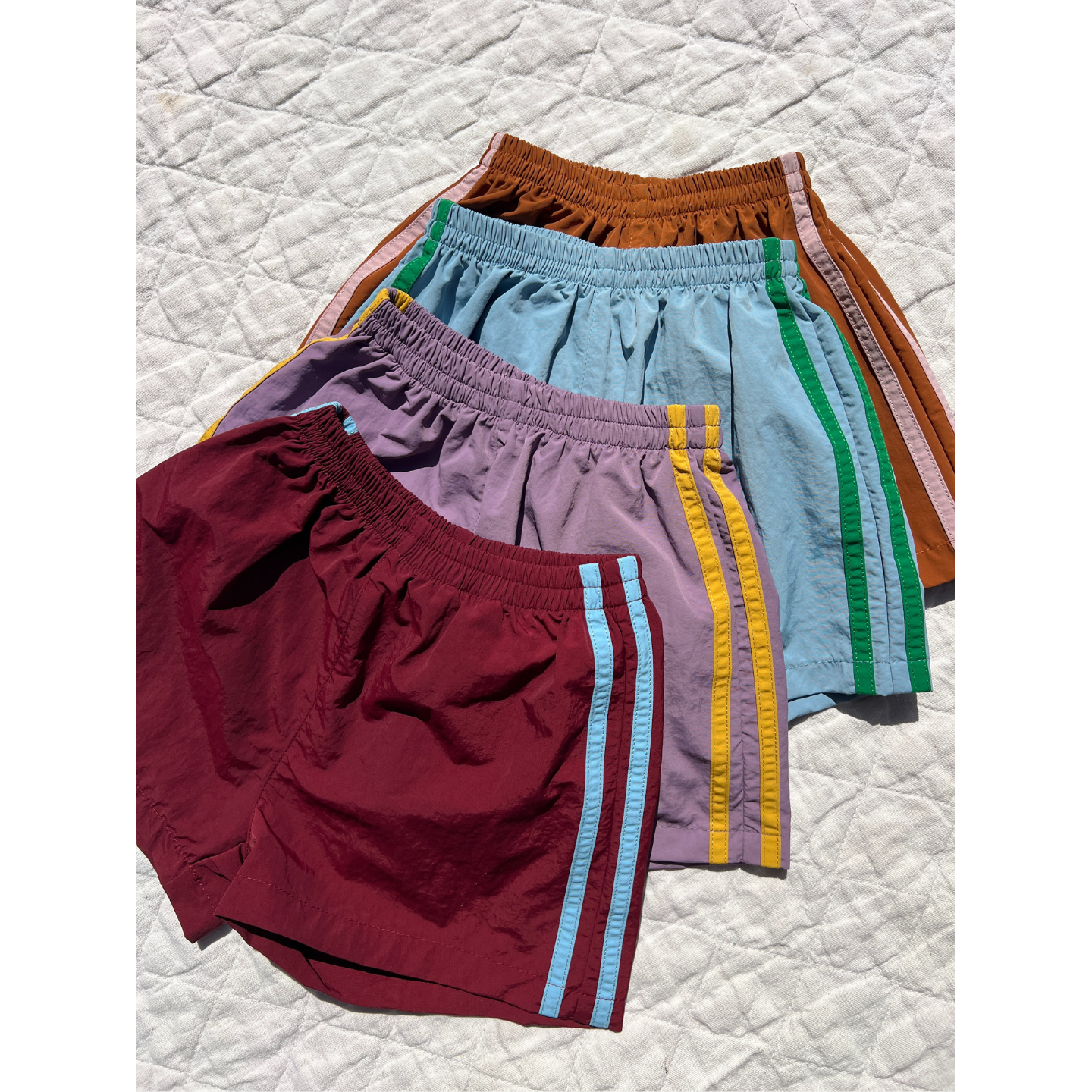 Maroon | Four pairs of SPRINTER SHORTS in red, purple, light blue, and brown with colored stripes and elasticated waists are laid out overlapping on a white quilted surface.