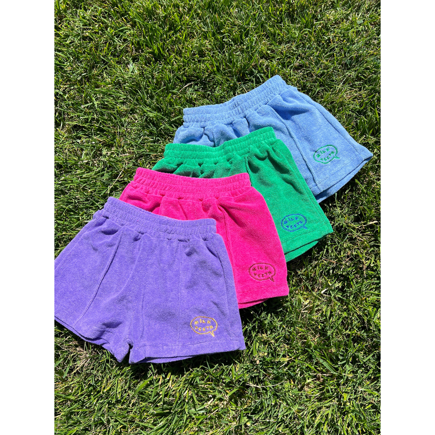Purple | A set of EVERYDAY TERRY SHORTS with elastic waists in vibrant summer colors is displayed on the grass, ideal for sunny days.