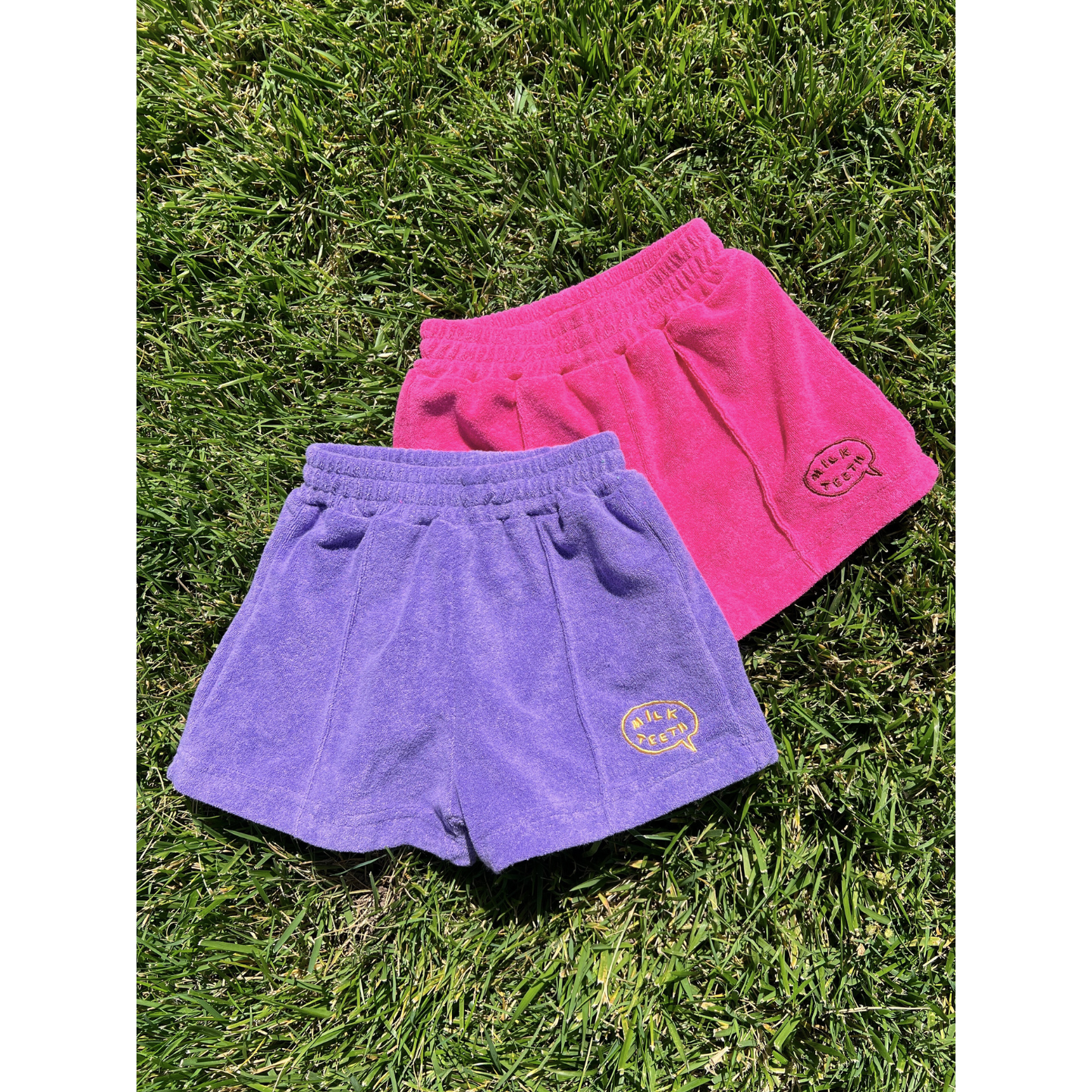 Purple | Two pairs of EVERYDAY TERRY SHORTS, one purple and one pink with yellow "MUK PEACH" logos and elasticated waistbands, are lying on green grass.