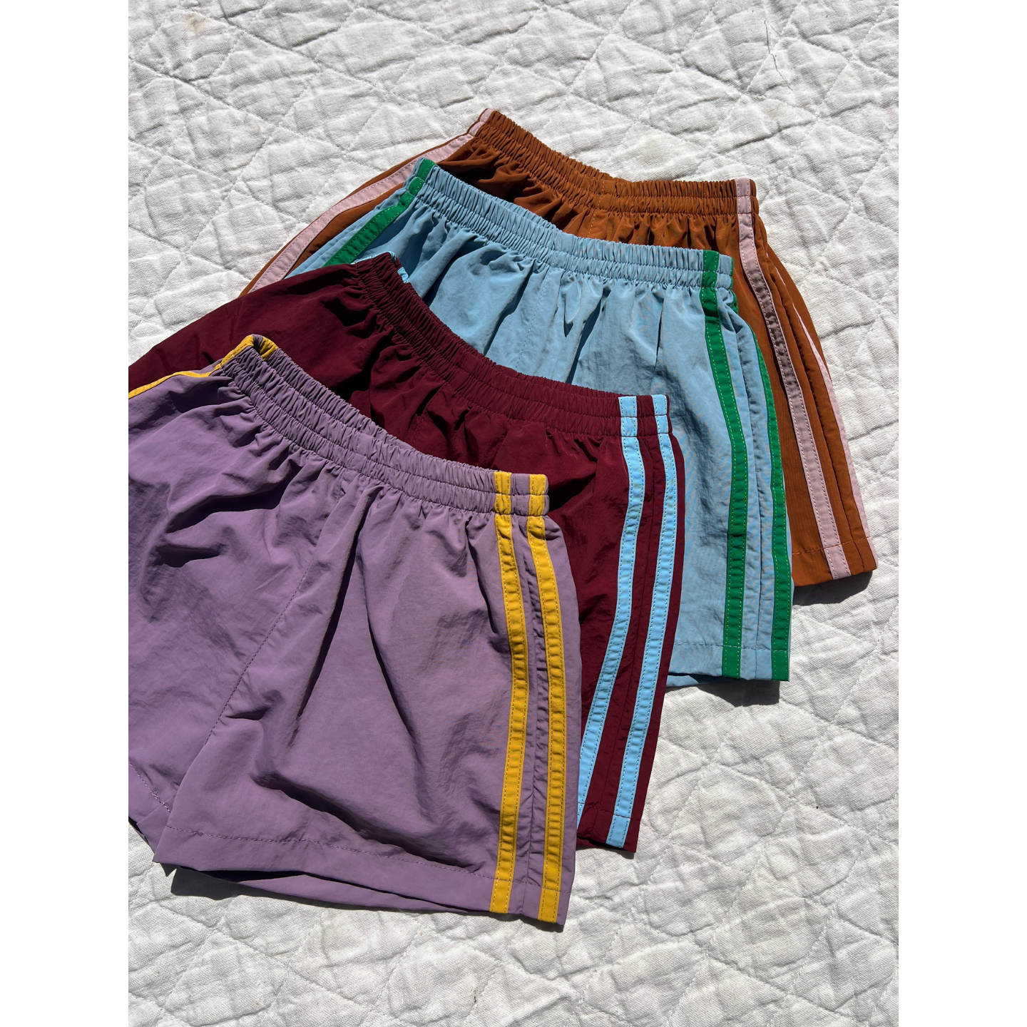 Purple | Four pairs of SPRINTER SHORTS with colorful stripes are arranged overlapping on a quilted white surface.