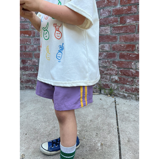 Purple | A child stands on concrete, wearing a white shirt with colorful faces, SPRINTER SHORTS with an elasticated waist, green socks, and blue sneakers. A brick wall is in the background.