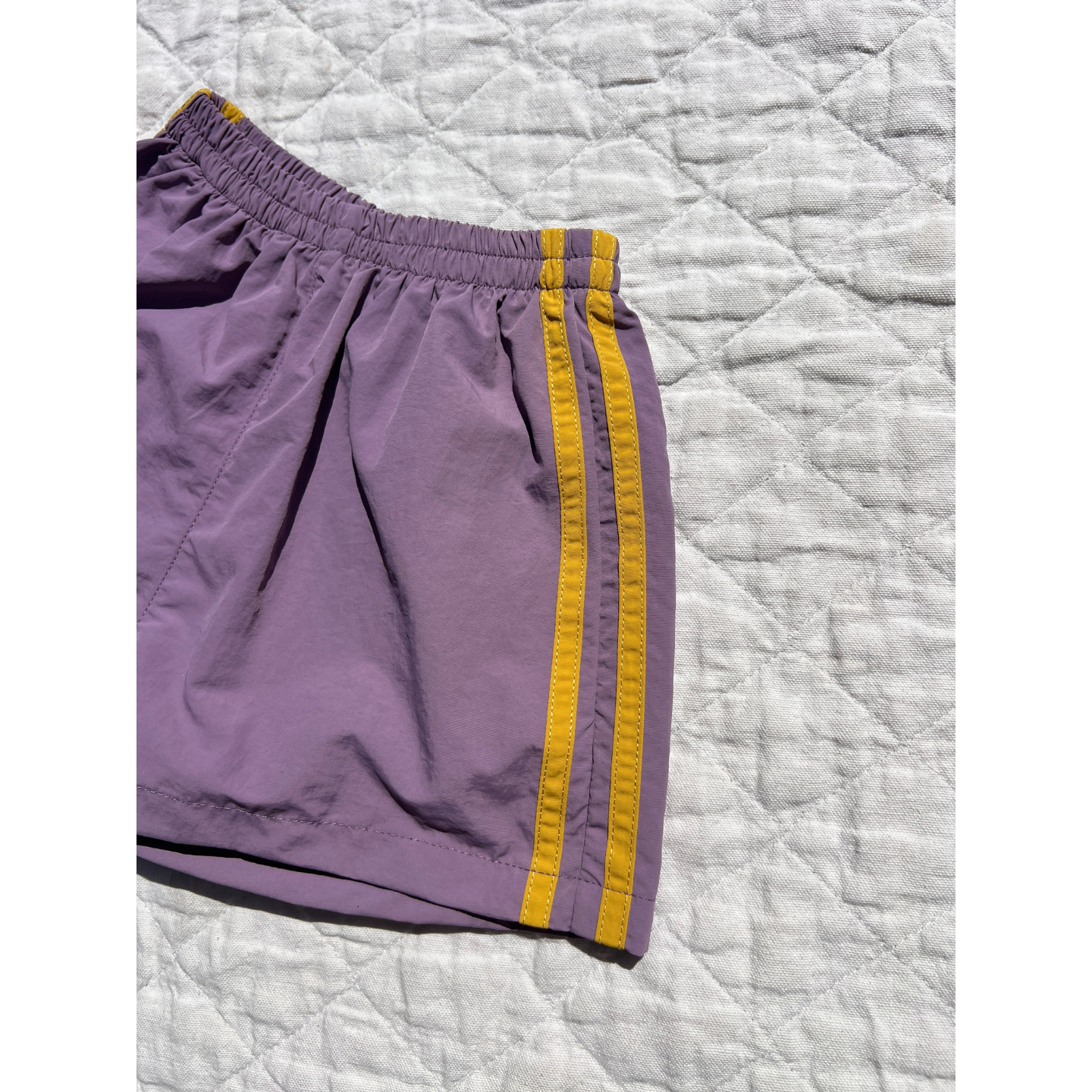 Purple | The SPRINTER SHORTS, featuring purple fabric, two yellow side stripes, and an elasticated waist, are displayed on a light quilted surface.