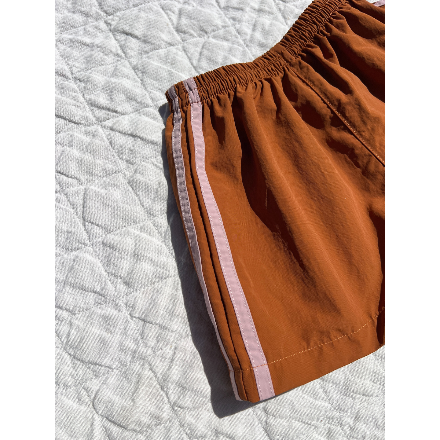 Rust | The SPRINTER SHORTS, featuring a vibrant orange color, light pink side stripe, and elasticated waist, are displayed on a textured white quilt in bright sunlight.