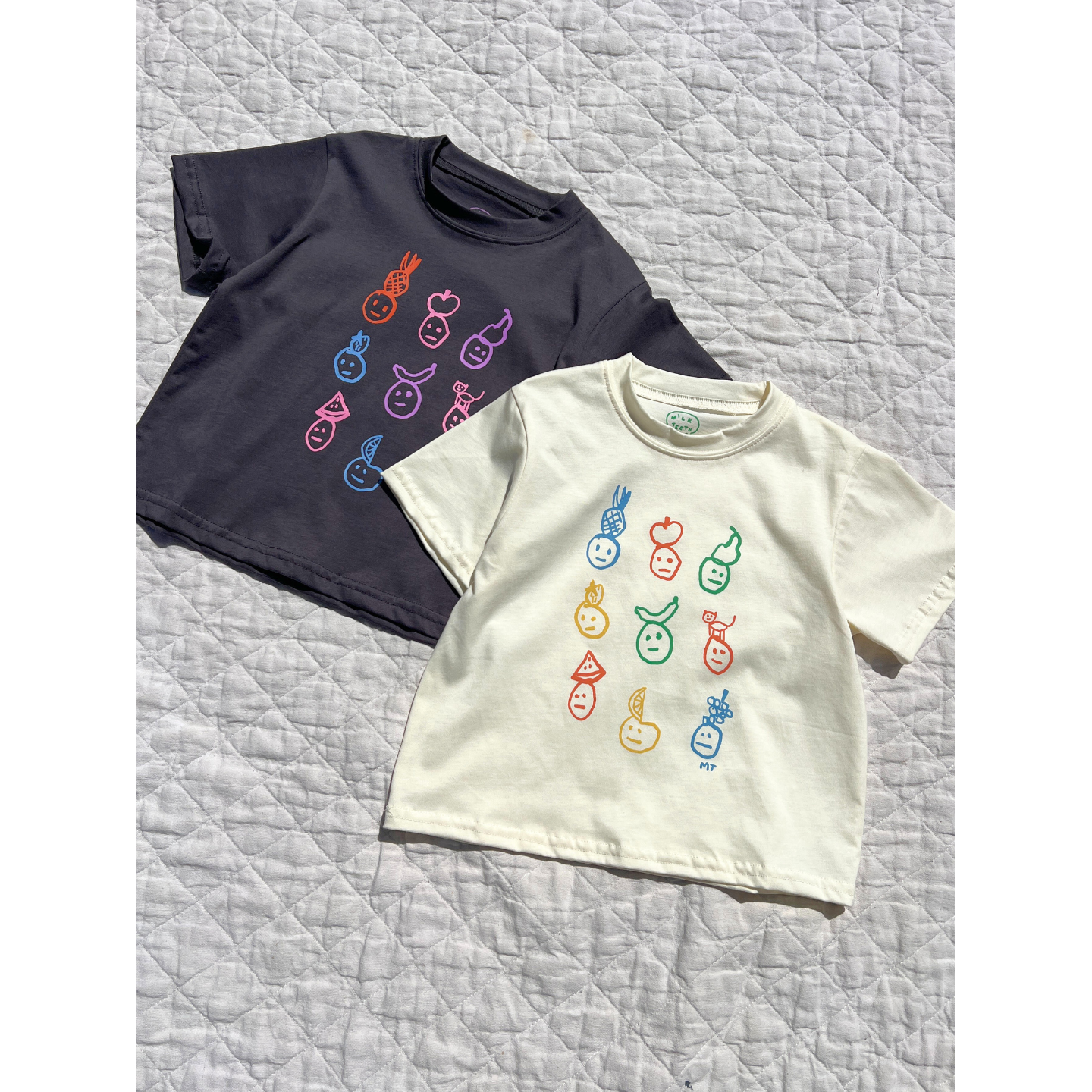 Ivory | Both colorwasy of the fruit face tee are laid flat on a quilt