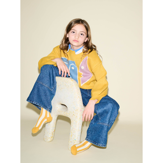 Marigold | A child with long hair sits on a uniquely shaped chair, wearing a comfy yellow sweater with a face design, blue jeans, and the STRIPE SOCK made of stretchy cotton. The background is plain beige.