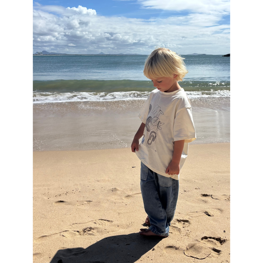 Medium Wash | Child standing on a sandy beach with ocean waves in the background