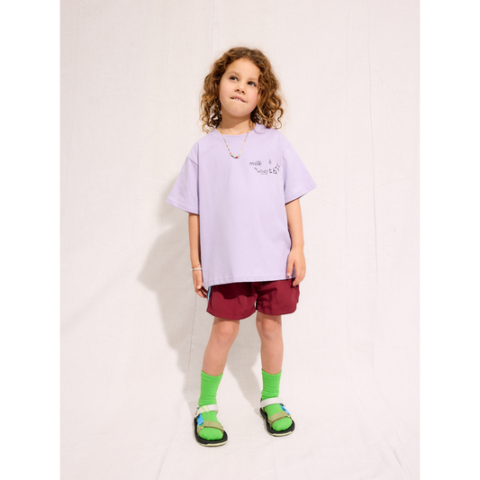 Purple | A child is wearing the purple Milk Teeth test with burgundy shorts and green socks and sandals