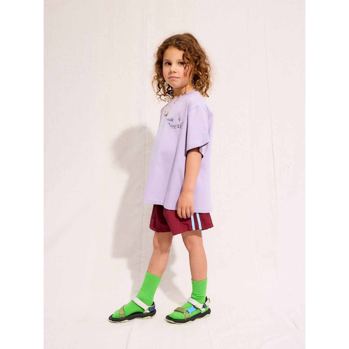 Purple | A child is wearing the purple Milk Teeth test with burgundy shorts and green socks and sandals