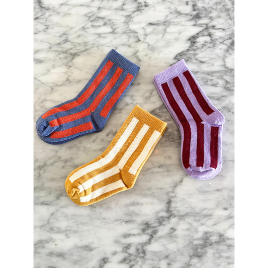Violet | Three pairs of STRIPE SOCK on a marble surface: one pair is blue with red stripes, one pair is yellow with white stripes, and one pair is lavender with maroon stripes. Made from stretchy cotton, these comfy socks are perfect for any occasion.