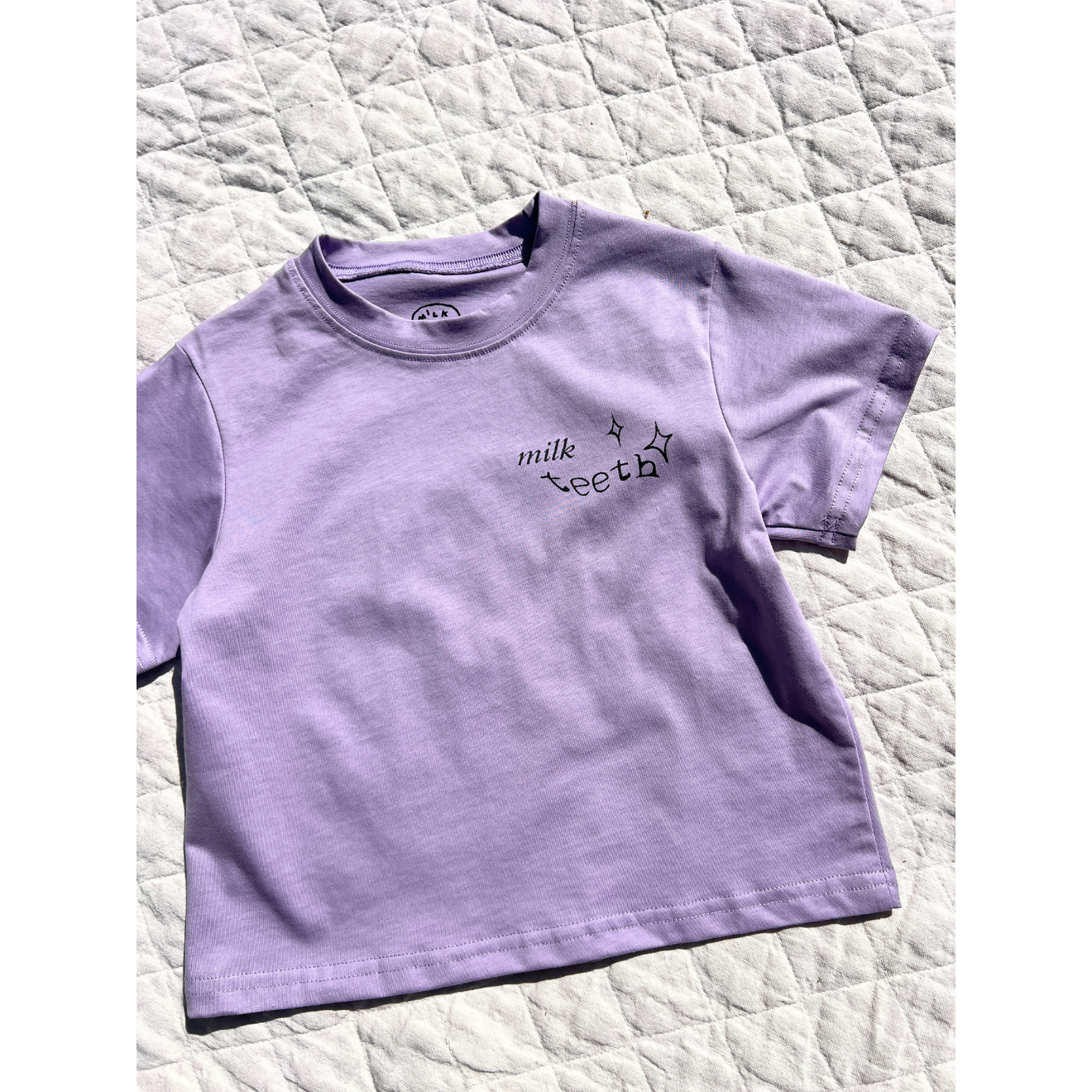 Purple | The MILK TEETH TEE, an artist-designed lavender short-sleeve shirt with black "milk teeth" text and star prints on the chest, is displayed on a quilted white surface.