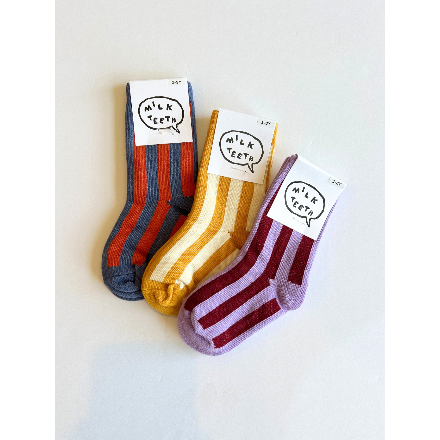 Slate | Three pairs of children's STRIPE SOCK are displayed with labels reading "Milk Teeth." Available in blue and red, yellow and white, and purple and red, these comfy socks are made from stretchy cotton, making them perfect for little feet.