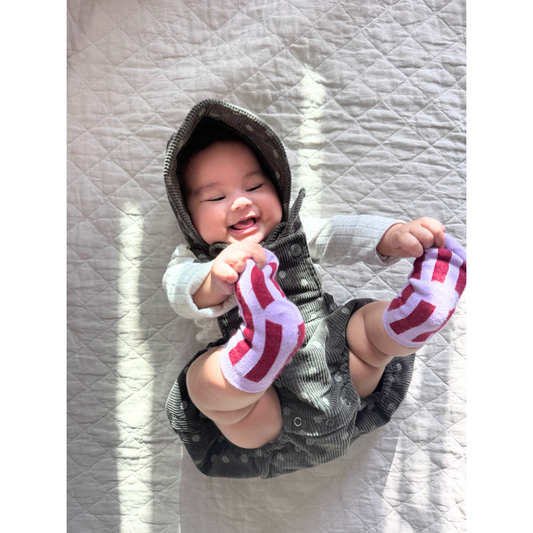 Violet | A smiling bag wears the STRIPE SOCKS: A pair of light violet, stretchy cotton socks featuring dark red vertical stripes. They wear a grey corduroy romper and matching bonnet, printed all over with polka dots. They are lying on a beige quilt and holding their toes.