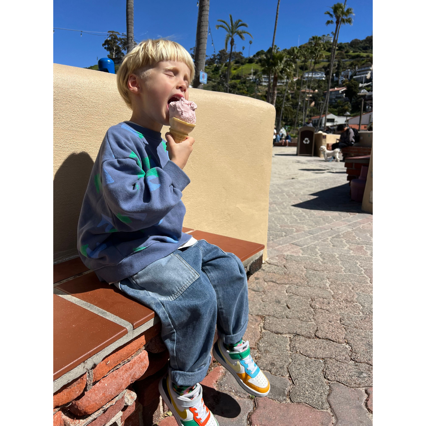 Medium Wash | A child wearing the double trouble kids jeans in medium blue denim with lighter blue patch pockets. They have short blond hair, and are wearing a blue sweatshirt and white sneakers with colorful trim, eating an ice cream cone on an outdoor bench with blue sky and palm trees.