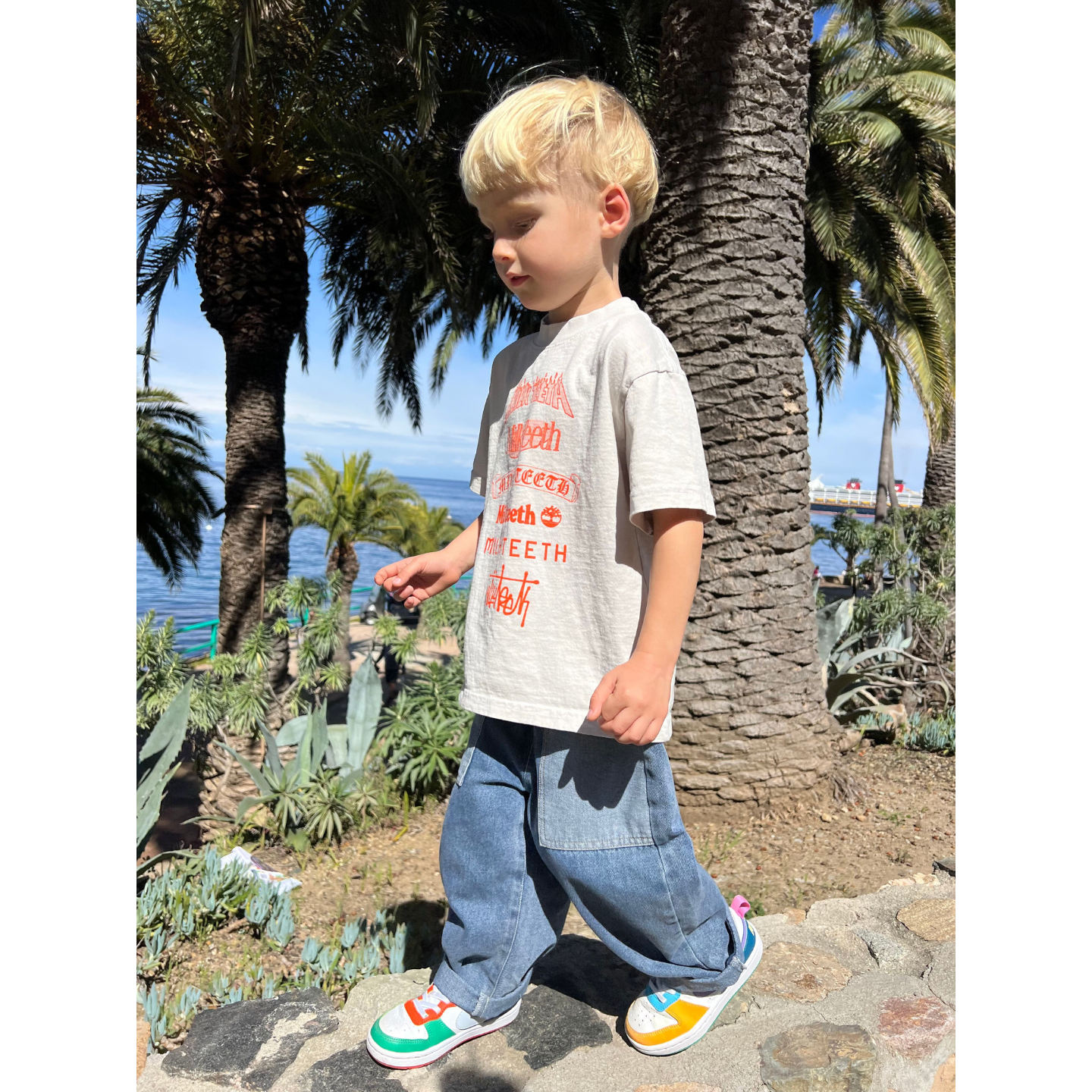 Medium Wash | A child wearing the double trouble kids jeans in medium blue denim with lighter blue patch pockets. They have short blond hair, and are wearing a light grey tshirt with orange Milk teeth lettering, and white sneakers with colorful trim, walking on a stone wall with palm trees, the ocean and blue sky in the background.