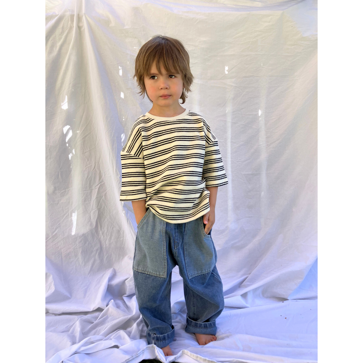 Medium Wash | A child wearing the Double Trouble baggy jeans in medium-blue denim with two large patch pockets of lighter denim. He wears an oversized striped t-shirt in cream, with black stripes, and stands in front of a white sheet.