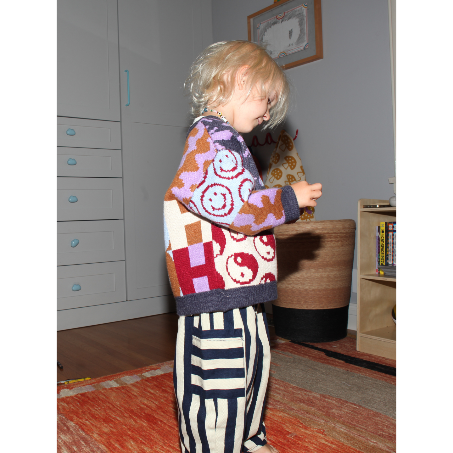 Child wearing a colorful patterned sweater and striped pants in a room with furniture and decor.
