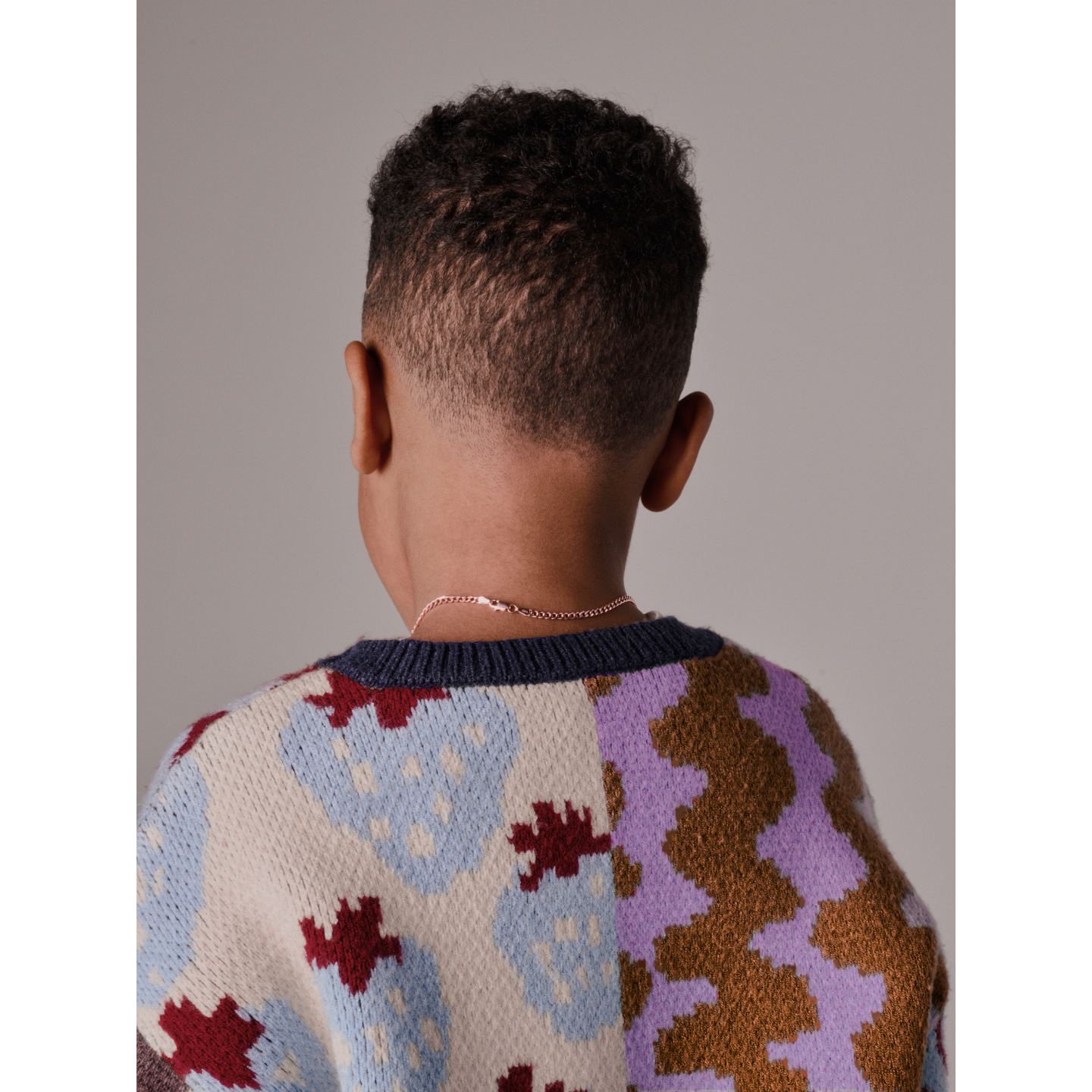 A child with short curly hair is seen from behind, wearing the EVERYTHING SWEATER featuring soft knit fabric with wavy checkers, paired with a thin chain necklace against a plain background.