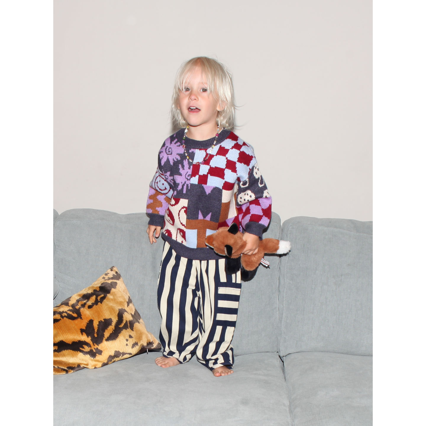 Child wearing a colorful sweater and striped pants holding a teddy bear on a couch.