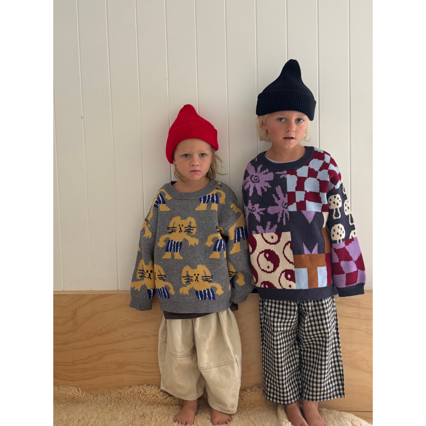 Gray | Two children wearing patterned sweaters and hats against a wooden floor and white wall background.