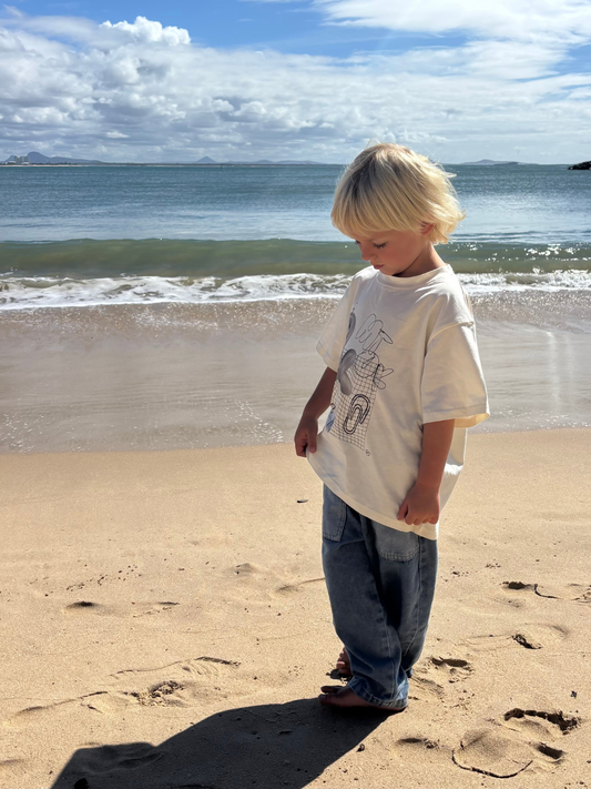 Medium Wash | Child standing on a sandy beach with ocean waves in the background
