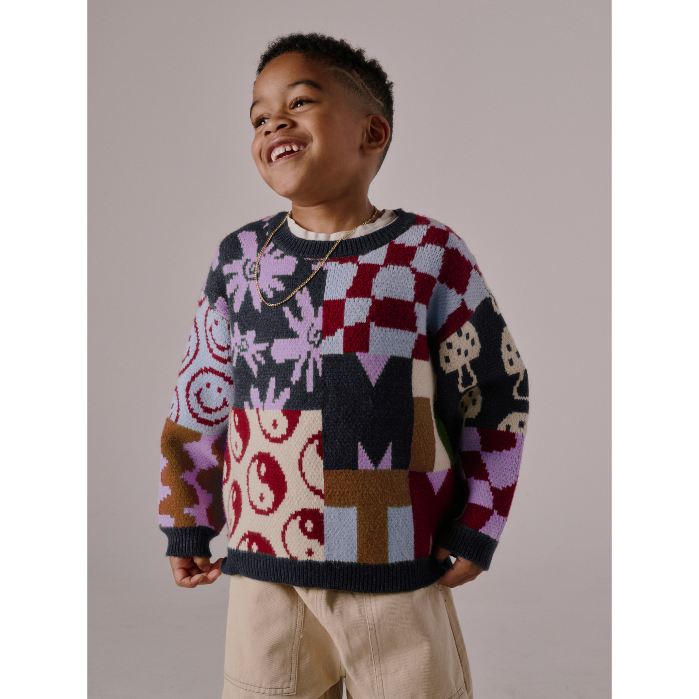 A young child smiles in the EVERYTHING SWEATER, featuring soft knit with wavy checkers and smiley faces, paired with light beige pants, standing against a plain background.